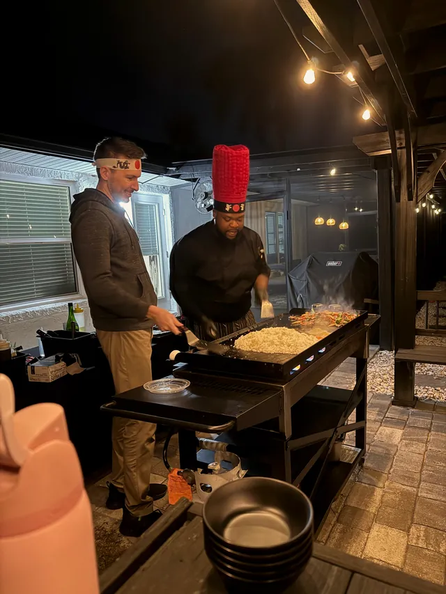 Chef cooking fried rice on a backyard grill while a guest watches beside him under string lights