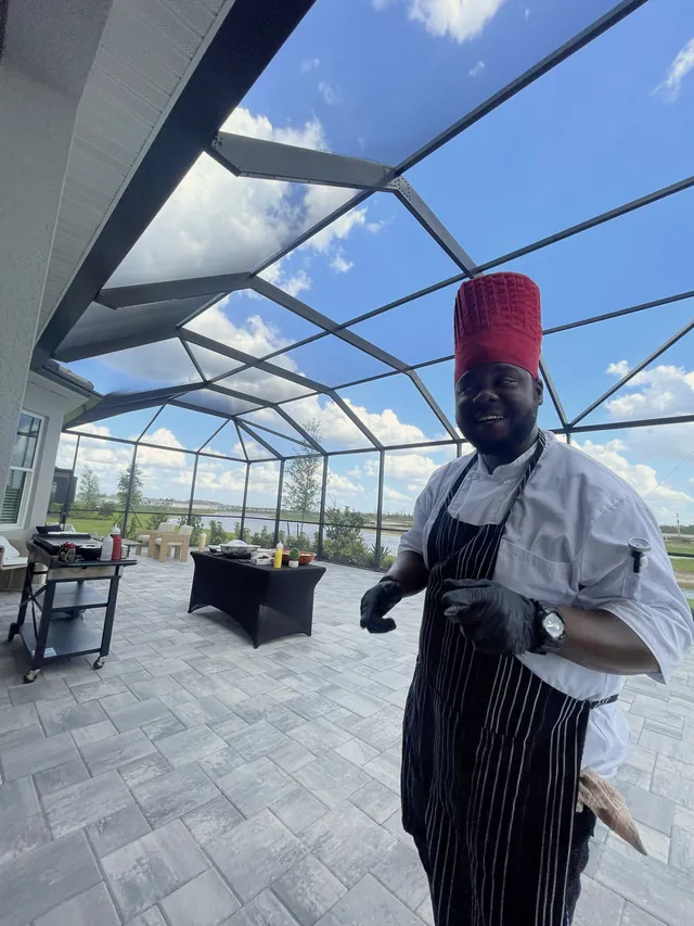 Smiling chef in a red hibachi hat and striped apron under a screened lanai